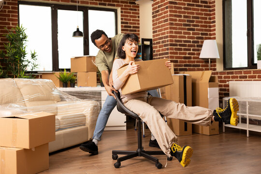 Smiling partners make memories on moving day, riding through living room with laughter and playful energy. Carefree couple laughing as black man pushes woman on chair through empty new apartment.