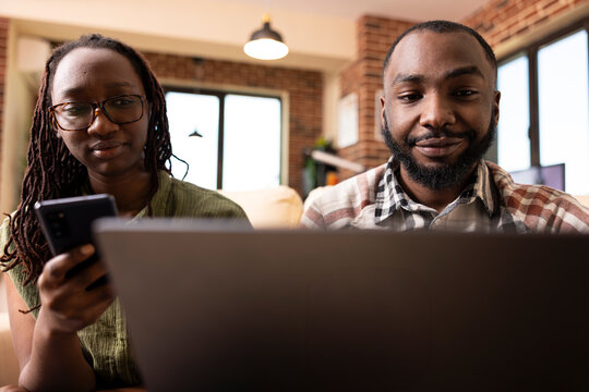 Close up of young black couple seated together using digital devices in modern brick wall room. Girlfriend chats on mobile phone while boyfriend works on laptop, showcasing modern tech lifestyle. - Powered by Adobe