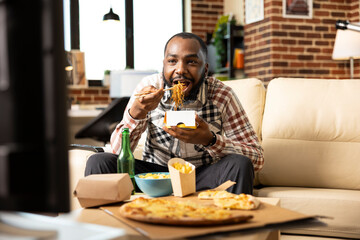 Black male individual eating noodles with chopsticks and binge watching his favorite show on tv in...