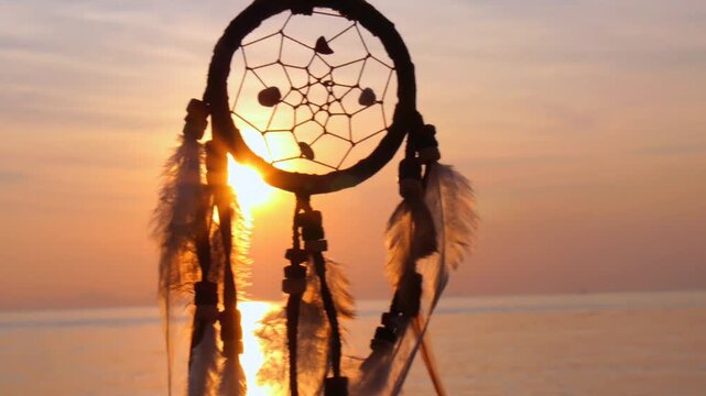 Silhouetted dreamcatcher with feathers gently swaying in the breeze against the background of a beautiful golden sunset over a calm sea, creating a peaceful and romantic evening atmosphere