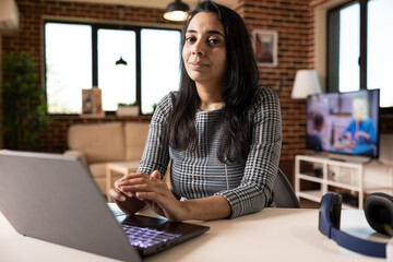 Confident female entrepreneur seated at home office desk with hands clasped, gazing at camera....