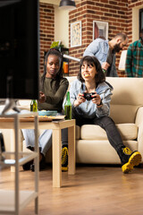 Caucasian woman with pizza slice in mouth, playing online multiplayer gaming with black female friend seated on couch. Multiethnic ladies enjoying video game during weekend hangout at home.
