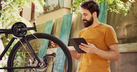 Healthy caucasian man multitasking inspecting and repairing his bike while utilizing his digital tablet for guidance. Young focused male cyclist browsing the internet to maintain modern bicycle.