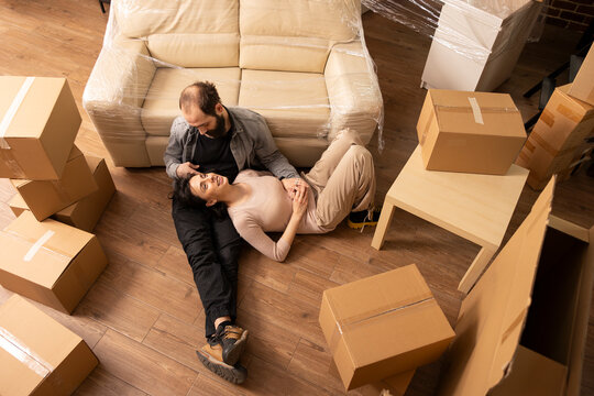 Young couple enjoy bonding on living room floor during moving day. Beautiful woman rests on lap of partner, surrounded by furniture and cardboard boxes, embracing relocation and new home ownership.