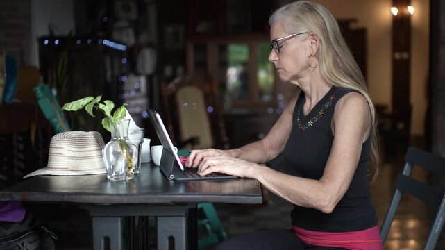 Mature woman sits at a cafe, sipping coffee and focused on her tablet, combining work and relaxation in a peaceful atmosphere.