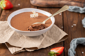 Rich and airy chocolate mousse in a white bowl, a golden spoon lifting a portion, garnished with delicate white flowers, resting on crumpled parchment paper and a wooden table