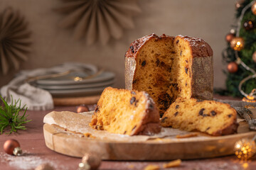 A sliced Panettone on a wooden cutting board with powdered sugar, set on a rustic table amidst holiday ornaments, stacked plates, and a decorated Christmas tree