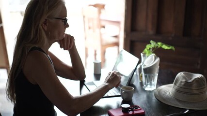 Mature woman focuses on her tablet while sipping coffee at a cafe, embracing the digital nomad lifestyle during the day.