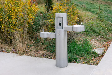 Modern stainless steel water fountain in a park surrounded by green plants and trees during a sunny...