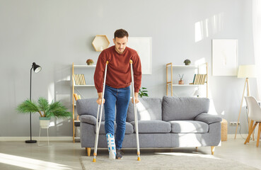 Injured young man with bandaged foot using crutches to move around modern living room. Casual dressed guy wearing orthopedic boot, recovering from leg injury, carefully walking during rehabilitation.