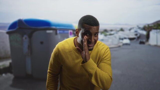 Black man holding lemon and cupping hand to mouth while smiling on a street near a dumpster and boats; playful secret.