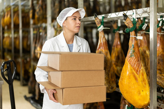 Employee of the jamon factory in a work uniform carries a stack of cardboard boxes. Young specialist prepares a batch of jamon to be sent in boxes to stores - Powered by Adobe