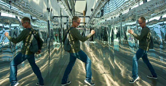 Infinite Reflections. A man poses playfully inside a long mirrored corridor, creating multiple distorted reflections along the chrome-like walls. Strong leading lines and reflective surfaces produce a