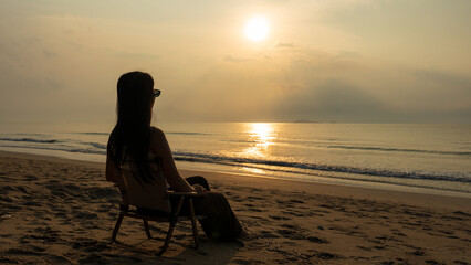 Woman relaxing on a beach chair facing the sunrise and the gentle waves.