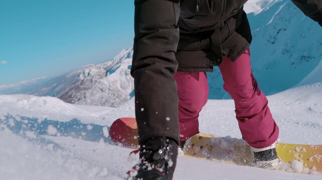 SLOW MOTION, CLOSE UP: Female snowboarder with boots strapped into a colourful board cuts through soft snow. Powder sprays around her legs with steep snowy mountains in the background on a sunny day.