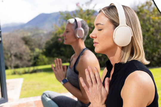 Two women meditating on mats wearing headphones following monitor on brick patio, copy space
