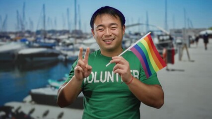 Young man holding rainbow flag at seaside port with volunteer shirt, signifying lgbt pride and diversity in outdoor setting with asian cultural influence. - Powered by Adobe