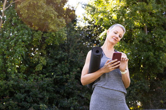 Senior woman holding yoga mat in sunlit park as checking fitness tracker and smartphone, copy space