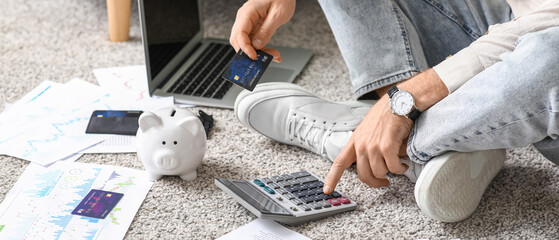 Young man in debt with credit card using calculator on carpet at home, closeup