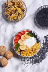 yogurt griego decorado con granola bananas frutillas arándanos y hojas de menta en fondo de mármol blanco ademas tiene nueces y semillas de sésamo negro