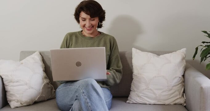 Woman settling onto gray sofa at home placing silver laptop on cross legs scrolling surprising feed
