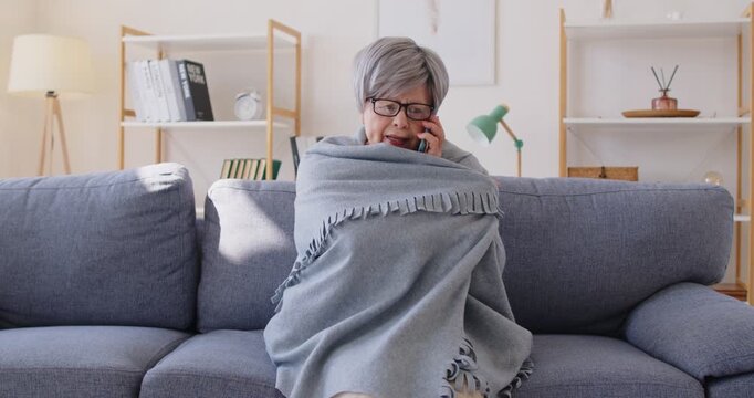 Elderly woman freezing in chilly room with low temperature calling public utility service by mobile phone. Senior person keeping warm under blanket sitting on sofa at home. Heating problems. 4k video