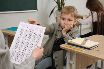 Worried schoolboy receiving answer sheet with C grade from teacher in classroom