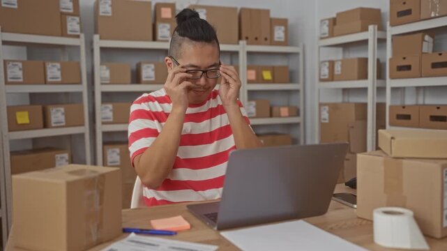 Man puts on glasses and types on laptop at a packing desk surrounded by stacked parcels and shelving in building; focused productivity.