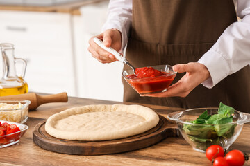 Woman spreading tomato paste on dough for pizza near table in kitchen