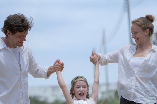 A joyful young family plays together outdoors under a bright sky. The loving parents swing their laughing daughter in the air, sharing a playful, happy moment on a summer holiday.