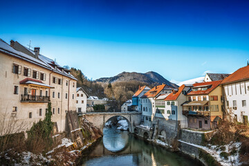 Winter panorama of Skofja Loka with the Selska Sora river and the Capuchin Bridge, also called Mamniti most. Stone arches, riverside houses and snowy details appear beneath a clear sky in Slovenia.