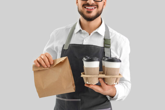 Young male barista with paper cups of coffee and food on grey background