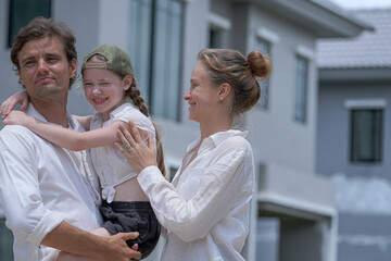 Fototapeta premium A happy Caucasian family, a father holding his daughter, and a mother, stand together outdoors. The man and child are smiling and pointing at something off-screen.