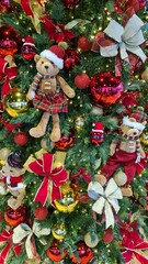 A richly decorated Christmas tree featuring plush teddy bears dressed in festive outfits, surrounded by large red and gold ornaments, glittery bows, poinsettias, and warm string lights. The tree is vi