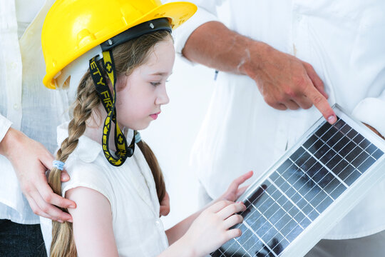 A curious young girl in a yellow hard hat learns about renewable energy. An adult teaches her about a solar panel, sparking interest in sustainability and future green technology.