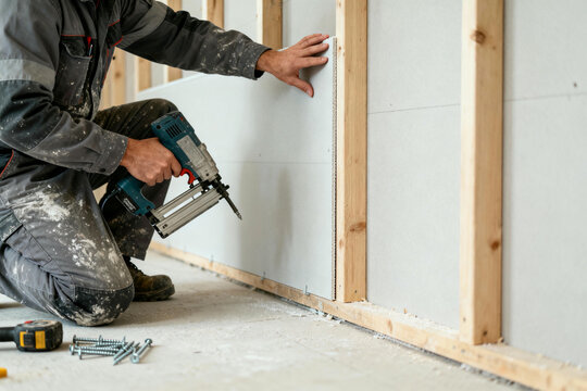 A skilled builder focuses intensely, fastening plasterboard with a power nail gun, as part of his meticulous construction project.