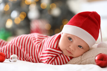 Cute little baby in pajamas and Santa hat with Christmas balls lying on bed at home