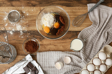 Bowl with ingredients for preparing tasty chocolate cake and baking utensils on wooden table in kitchen, top view