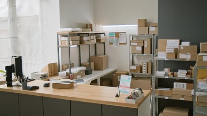 Full no people shot of interior of courier delivery and logistics office, with customer service counter with computer and advertisement leaflet, parcels and packages stacked on racks in background