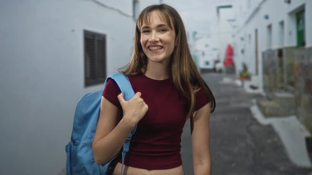 Young woman holds backpack strap on a narrow city street, lips pursed in a small grin while wearing burgundy crop top; playful.