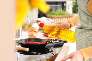 Housewife pouring sunflower oil into frying pan with hot chili peppers in kitchen, closeup