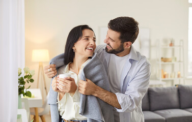 Smiling caring young husband gently covering his cheerful wife holding cup of tea with warm plaid. Happy young family couple standing together by window, hugging and enjoying winter time at home.
