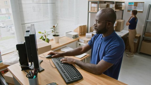 Medium shot of busy, focused young African American express delivery company employee standing behind counter in office, working on computer, preparing consignment forms for parcels