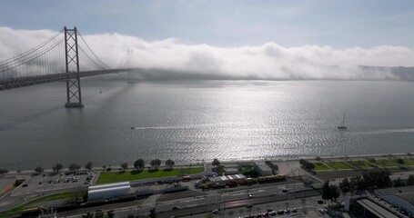 Aerial drone shot towards the 25th April Bridge and waterfront in Lisbon, Portugal, Europe. Sunny and bright with clouds, fog, mist covering the bridge. Shot in ProRes 422 HQ