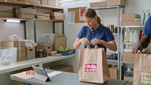 Medium shot of young Caucasian female dark store associate stapling customer order invoice to paper bag with groceries, ready for delivery, African American colleague collecting produce in background