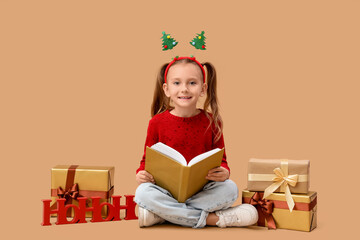 Naklejka premium Cute little girl with book and Christmas gifts sitting on beige background