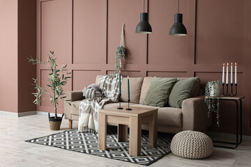 Interior of living room with sofa, houseplants, nightstand and candle holders on coffee table near brown wall