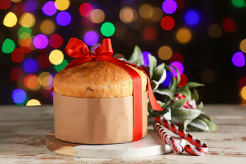 Tasty Christmas Panettone with red bow and decorations on white wooden table against blurred colorful garland background
