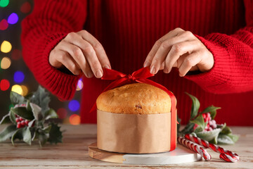 Woman with decorations tying red bow on tasty Christmas Panettone at wooden table near Christmas tree, closeup