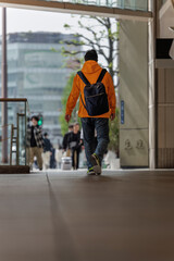 Casually dressed man in bright orange hoodie walks through corridor carrying dark blue backpack slung low in a relaxed manner on his back.